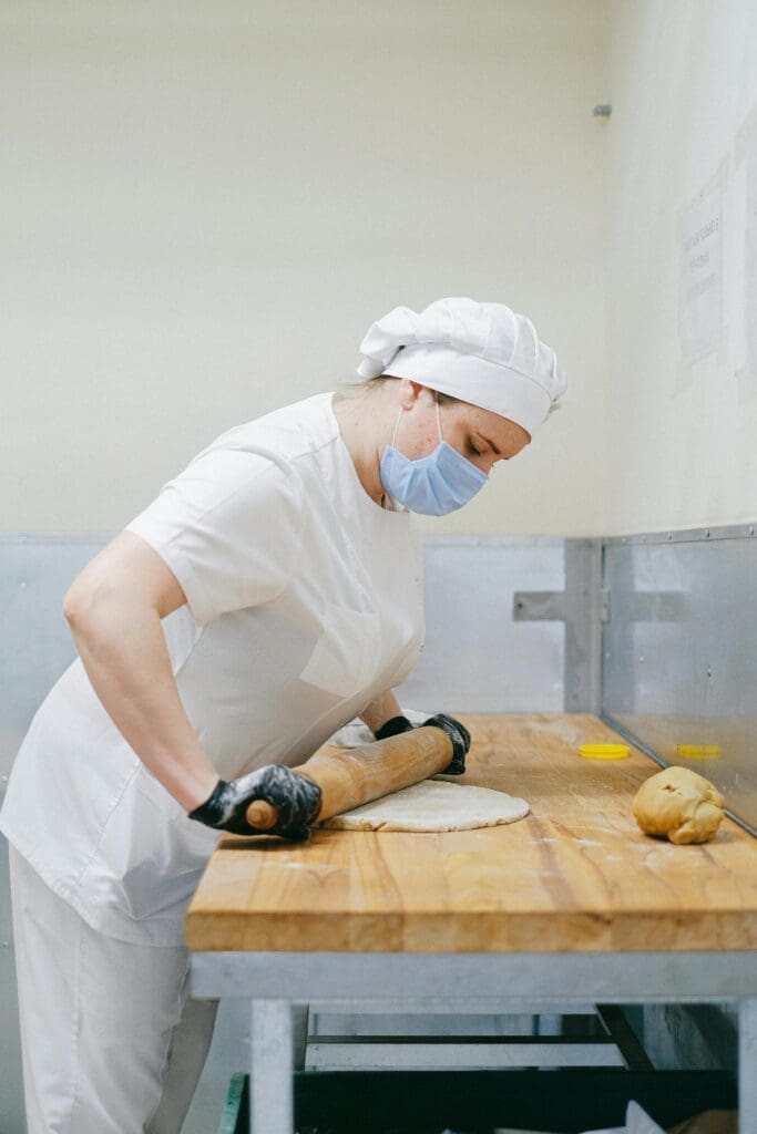 pexels-photo-5964528-5964528 A masked pastry chef uses a rolling pin to flatten dough on a wooden surface in a kitchen.